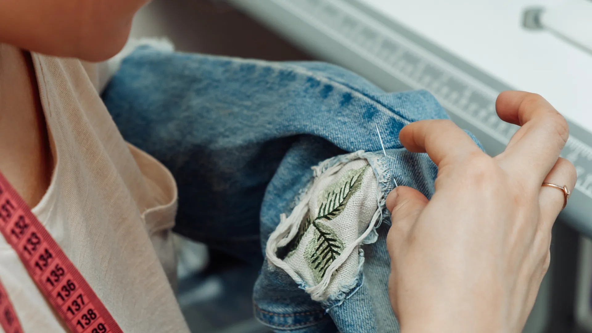 Eine Person repariert eine Jeans mit Visible Mending, indem sie ein Upcycling-Patch mit Pflanzenmotiv anbringt.