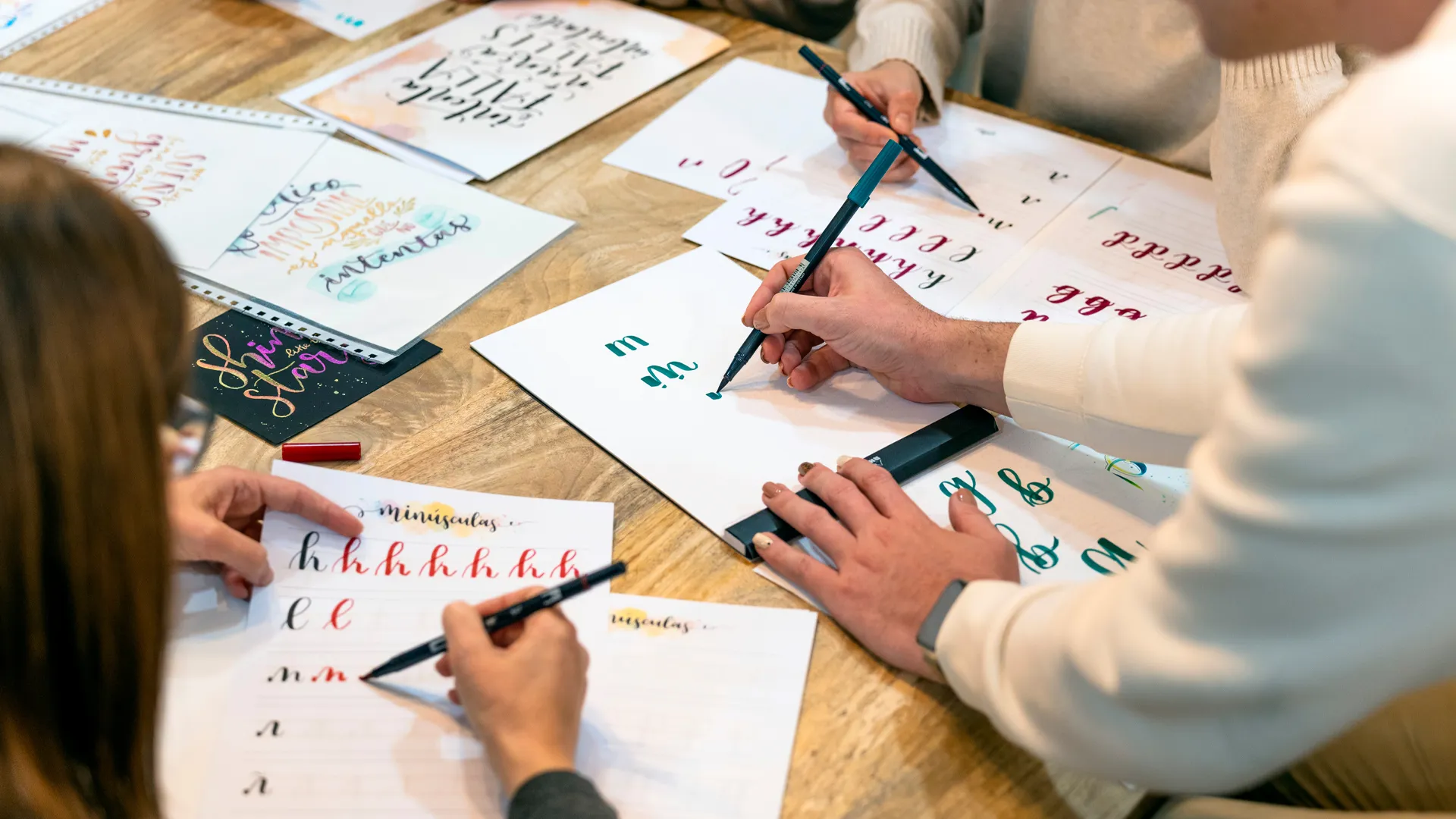 Mehrere Personen üben Handlettering mit Brush Pens. Auf dem Tisch liegen Vorlagen, bunte Schriftproben und Übungsblätter.
