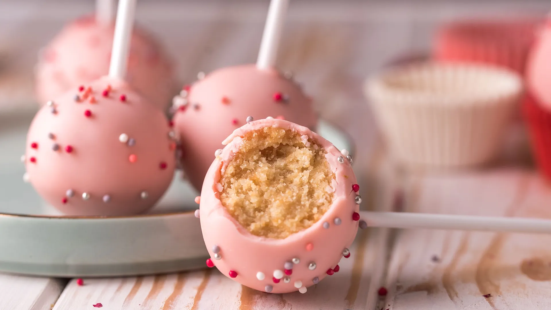 Ein angebissener rosa Cake Pop mit Streuseln liegt auf einem Teller, umgeben von weiteren Cake Pops.