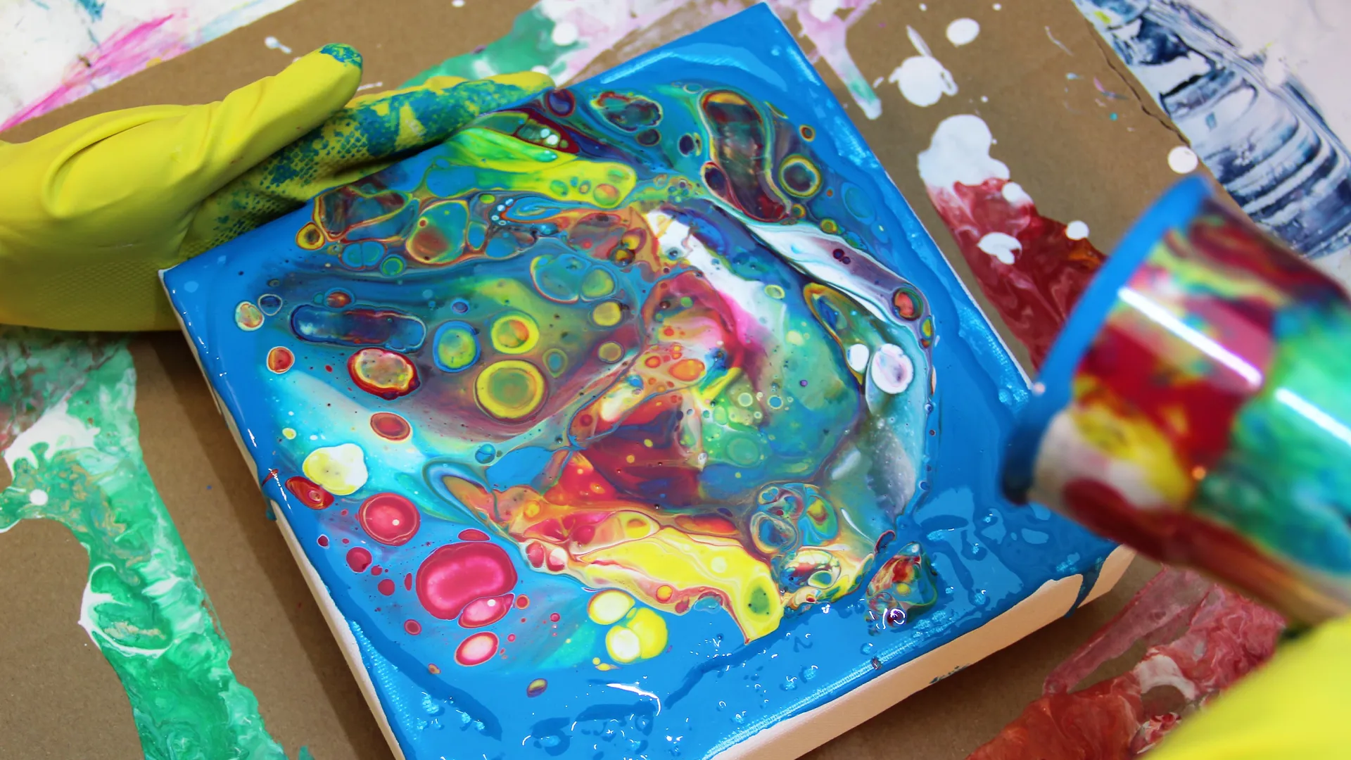 Eine Person mit Handschuhen giesst flüssige Farbe auf eine Leinwand. Diese Technik nennt sich Fluid Painting und erzeugt abstrakte Muster.