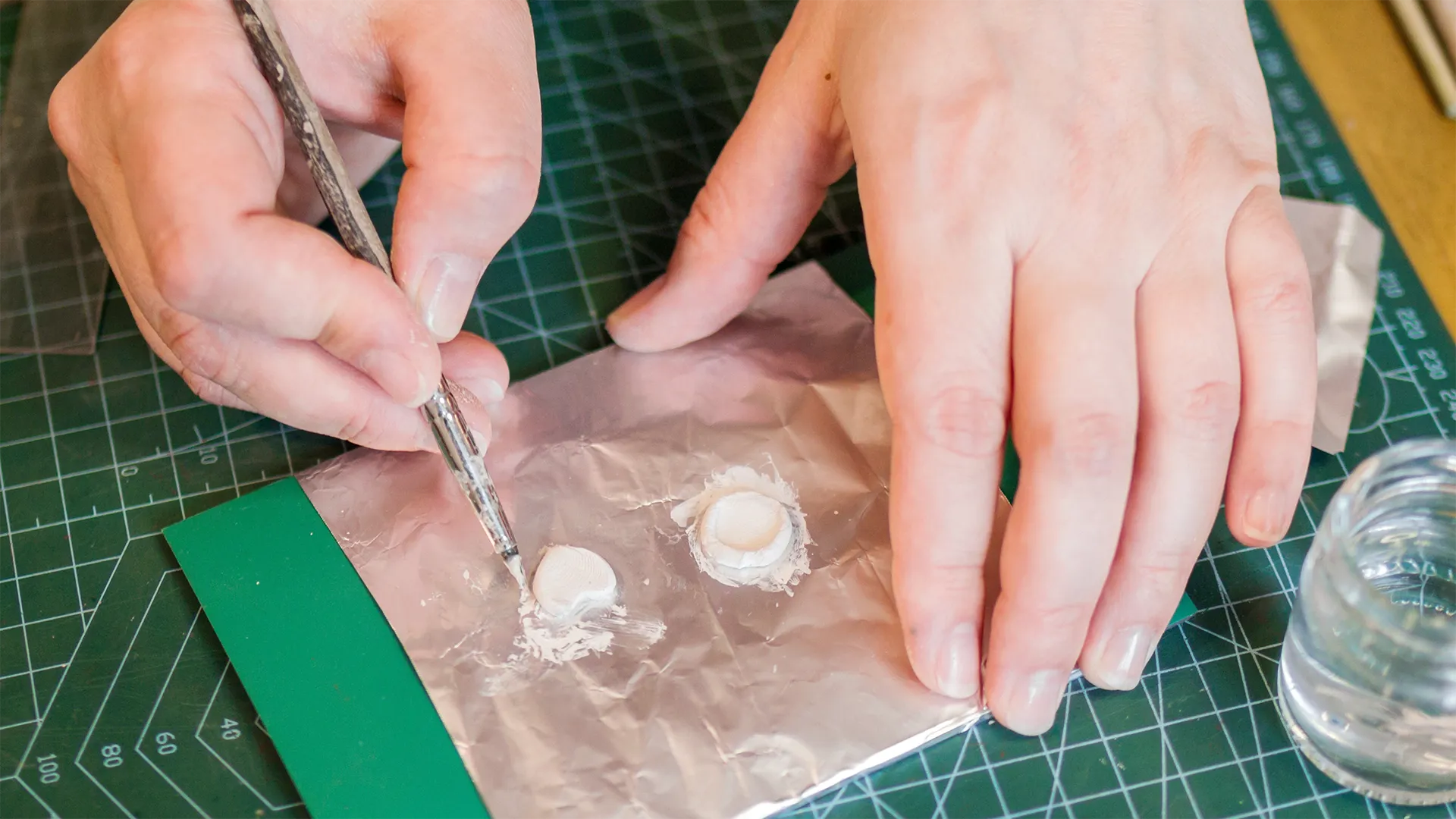 Hände formen mit einem Werkzeug kleine Objekte aus Art Clay Silver auf einer Unterlage aus Aluminiumfolie.