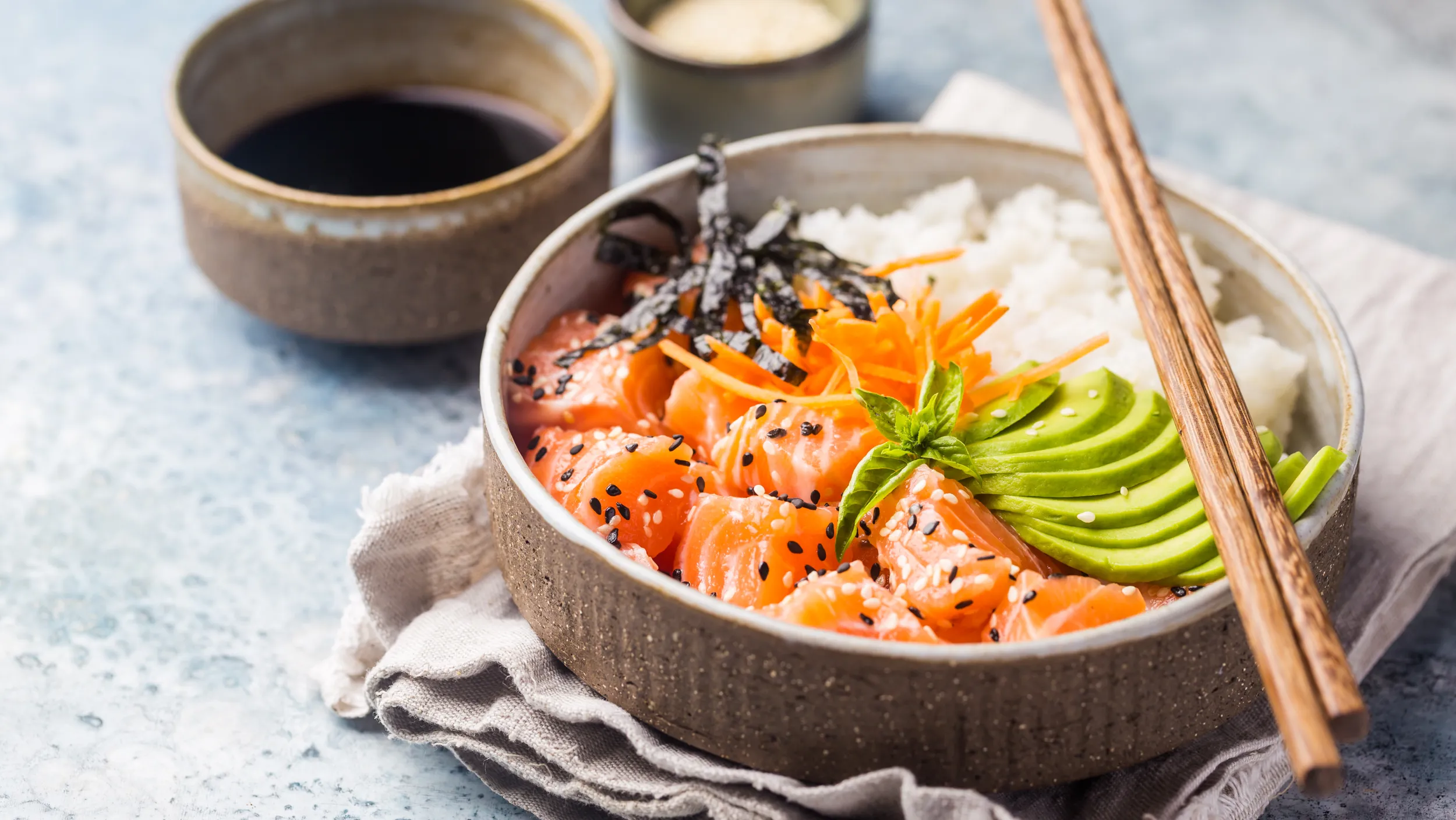 Eine Bowl mit Reis, Lachs und Avocado und Soyasauce serviert in einer Schale mit Stäbchen.