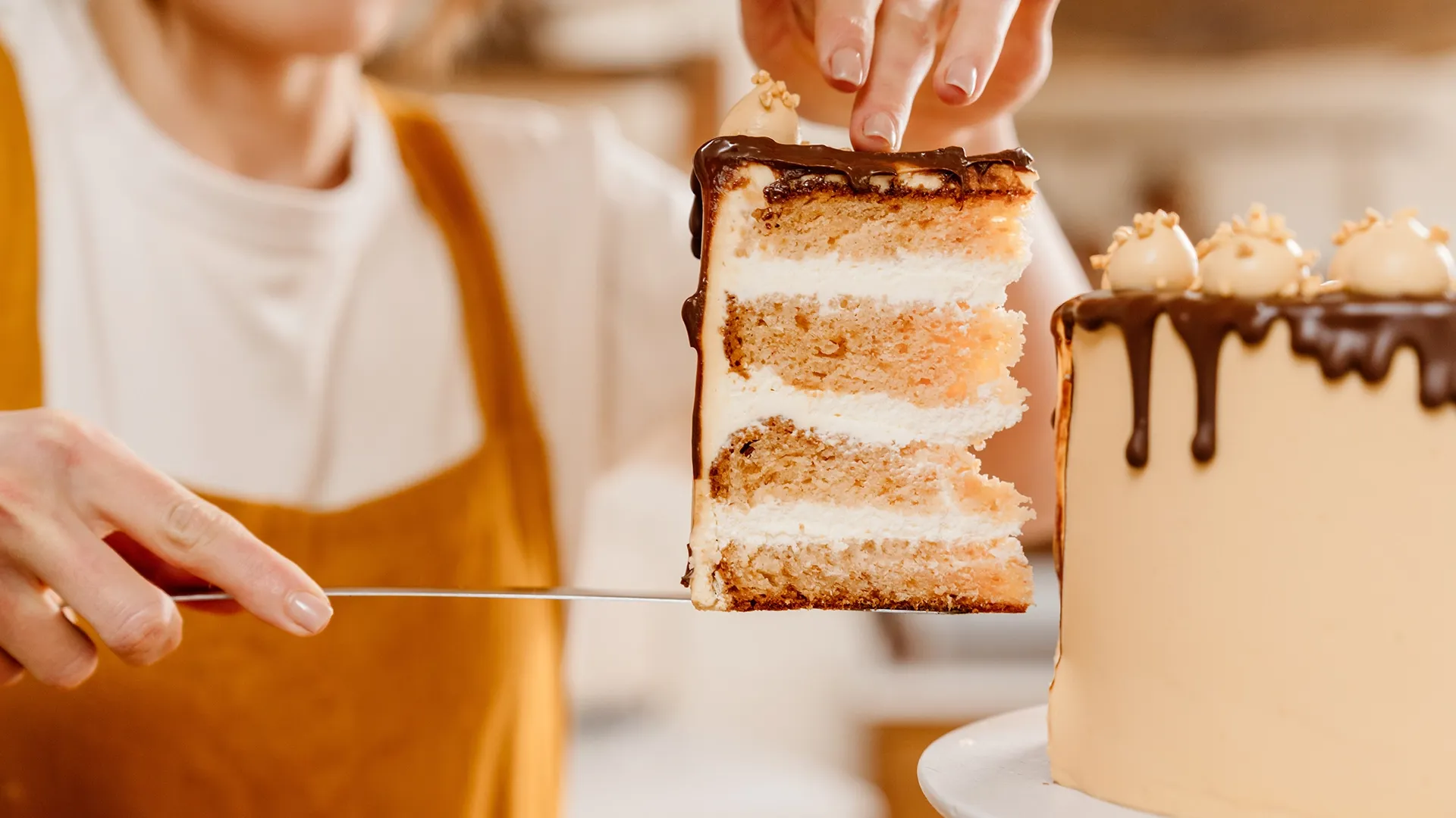 Ein Stück einer Torte mit Schokoladenglasur wird mit einem Messer serviert, während die restliche Torte auf einem Teller steht.