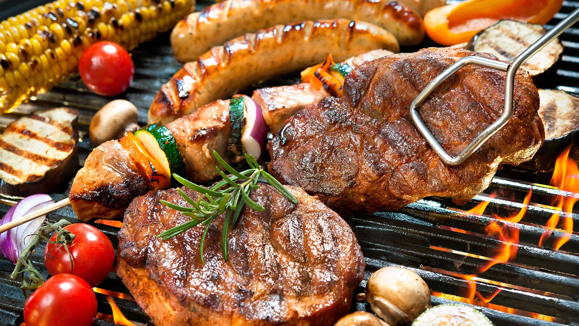 Verschiedene Grilladen liegen auf dem Grill. Die Grillzange greift nach dem Steak.
