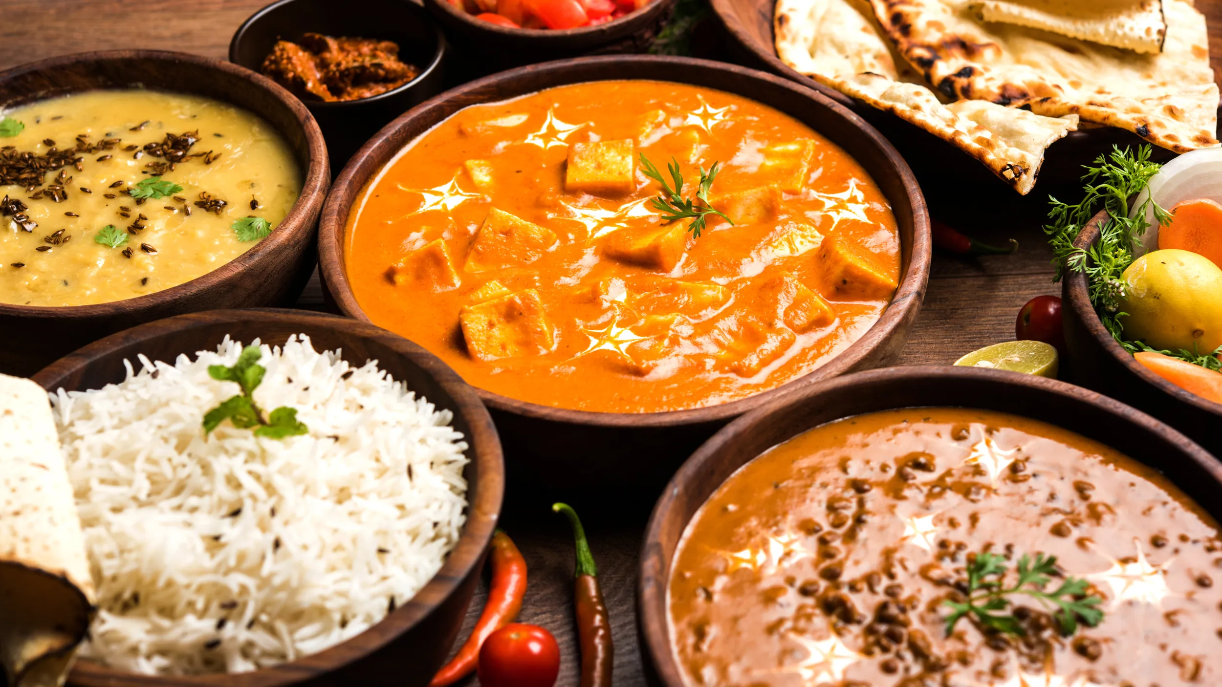 Eine Auswahl an Gerichten der indischen Küche: Dal, Paneer-Curry, Linsengericht, Basmatireis, Naan-Brot und verschiedene Beilagen wie Chutney, Tomaten und Zitrone, serviert in Holzschalen.