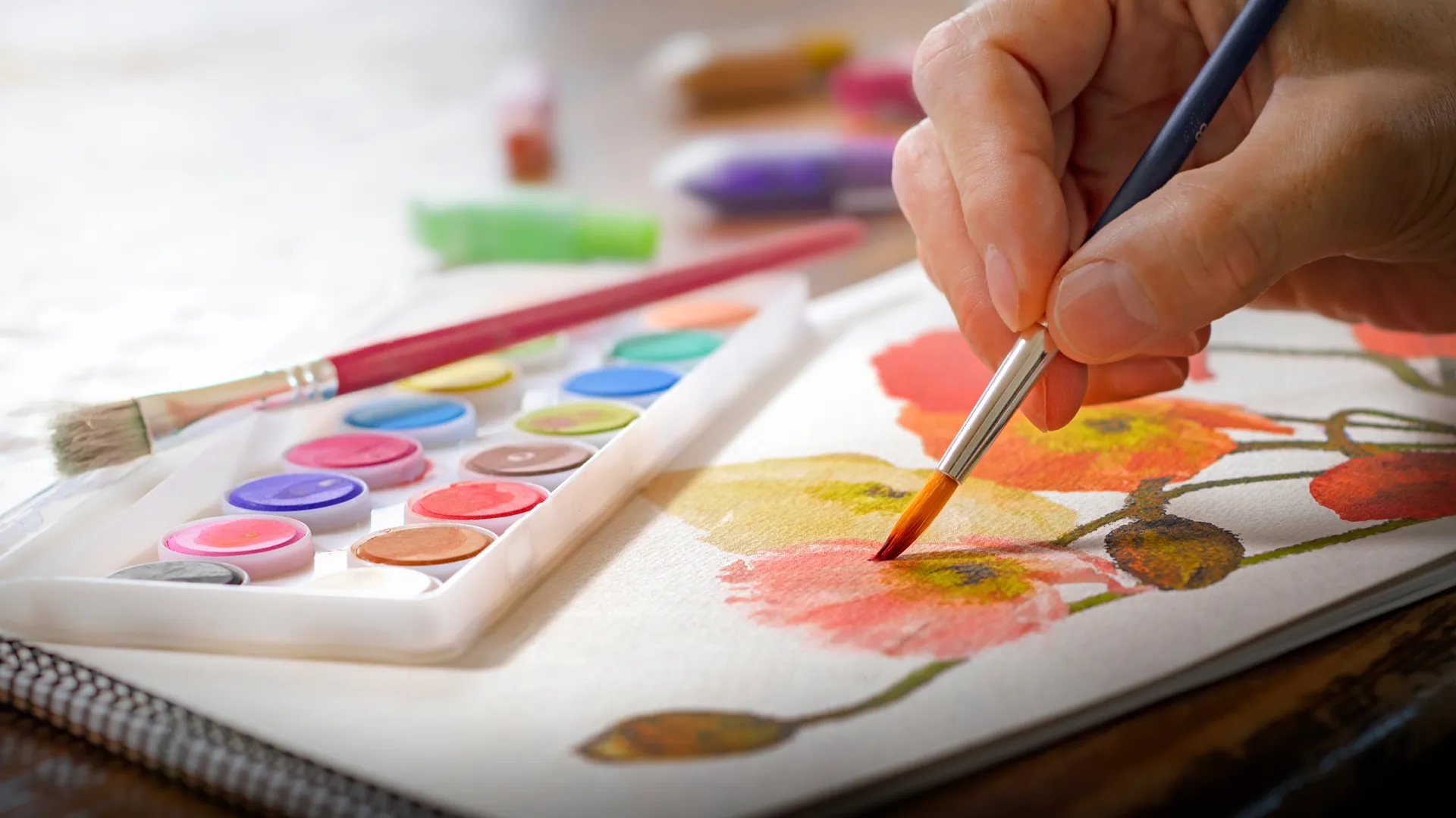 Eine Person malt mit Aquarellfarben Blumen auf Papier. Pinsel und sanfte Farbtöne schaffen ein zartes, künstlerisches Motiv.