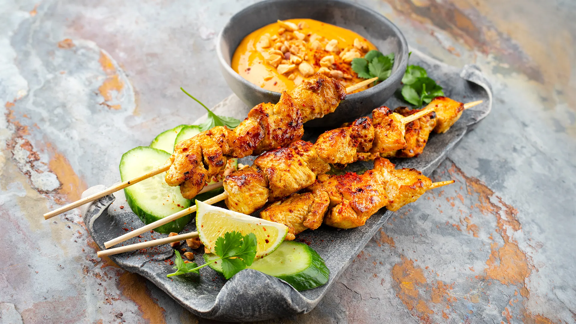 Thailändisches Fingerfood: Drei Satai-Spiesschen serviert auf einem Teller mit Erdnusssauce.